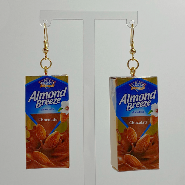 Gold hanging earrings with a special edition, gold, mini, chocolate Almond Breeze almond milks as the charm. The charms are small metallic gold and shiny finished cardboard boxes replicating boxed almond milk. The packaging of the almond milk has a blue backdrop with almonds falling into a pool of chocolate milk. Earrings are hanging on a clear acrylic earring holder with a plain white backdrop.