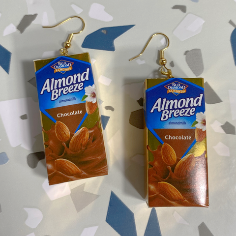 Gold hanging earrings with a special edition, gold, mini, chocolate Almond Breeze almond milks as the charm. The charms are small metallic gold and shiny finished cardboard boxes replicating boxed almond milk. The packaging of the almond milk has a blue backdrop with almonds falling into a pool of chocolate milk. Earrings are placed on a blue and grey terrazzo backdrop. 