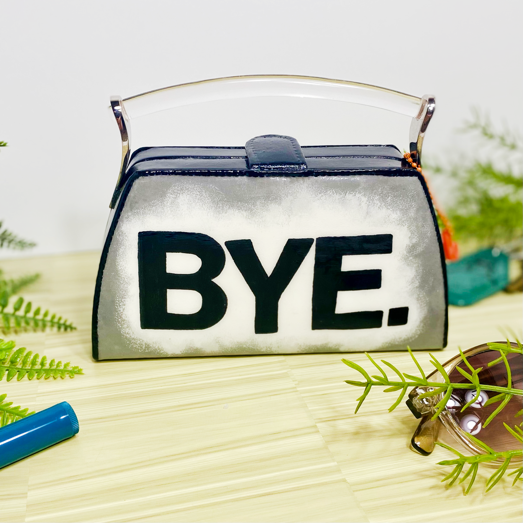 Box clutch with handpainted word, "bye", on a white background that fades into silver. Purse has clear handle and magnetic latch. Purse is on wood surrounded by faux plants.