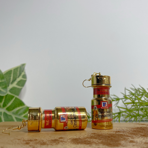 Mini gold McCormick cinnamon spice earrings on a wood cutting board with a white backdrop. There is cinnamon dusted in front of the earrings on the cutting board and faux plants in the background.