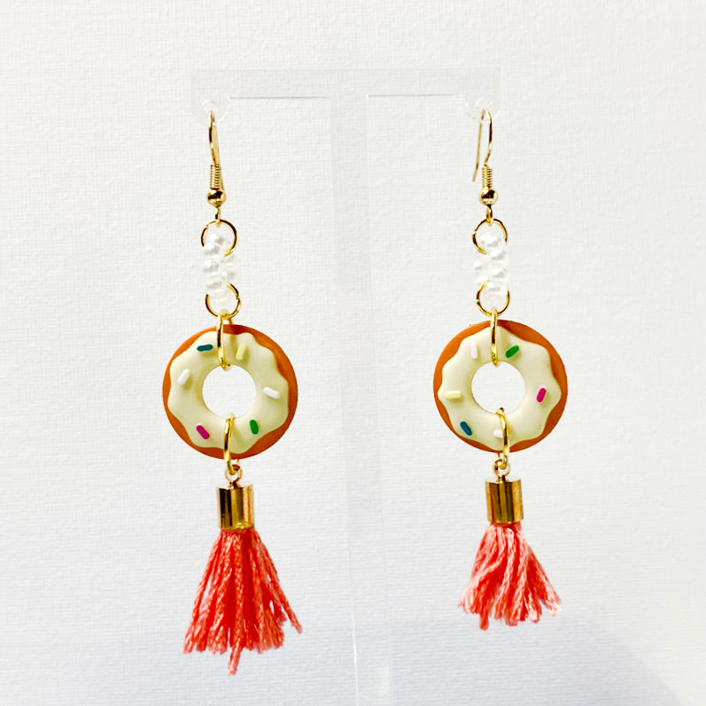 Gold-toned hangings earrings with an acrylic pearl ring, donut with yellow frosting, and an orange tassel.