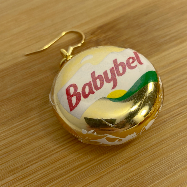 Mini gold Babybel cheese earring on a wood backdrop.