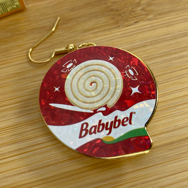 Mini gold Babybel swirl cheese earring on a wood backdrop.