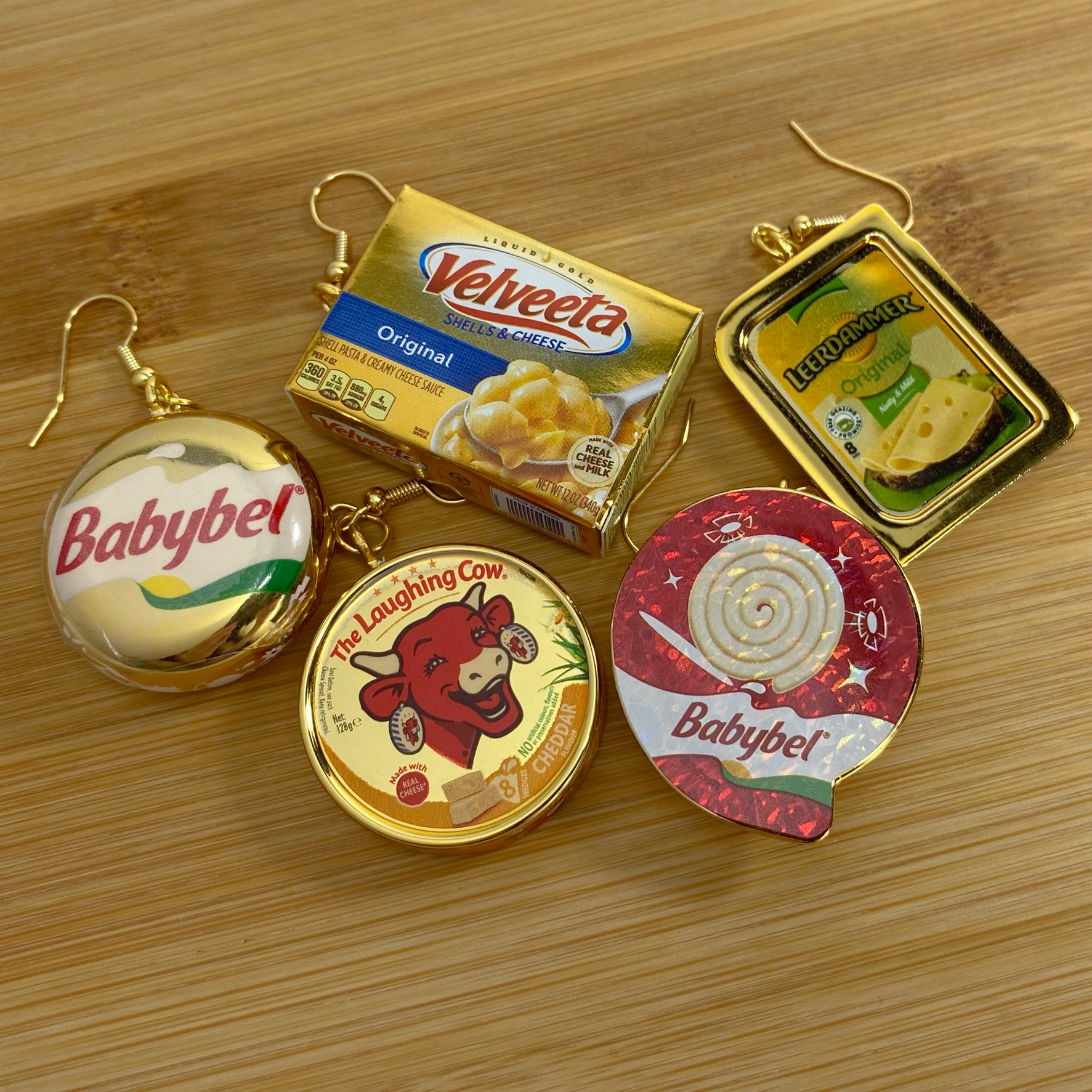 Mini gold Babybel, Velveeta, Babybel Swirl, Laughing Cow, and Leerdammer cheese earrings on a wood backdrop. 
