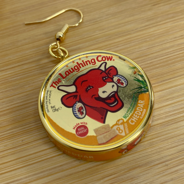 Mini gold Laughing Cow cheese earring on a wood backdrop.