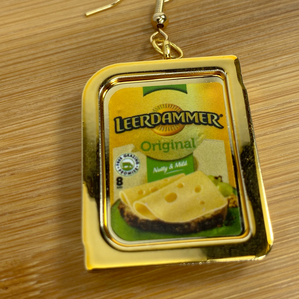 Mini gold Leerdammer cheese earring on a wood backdrop.