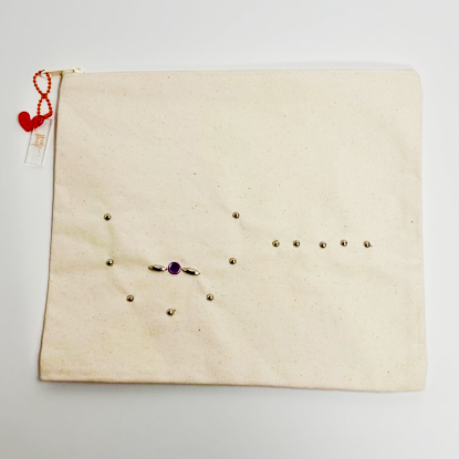 Canvas pouch bedazzled with silver metal studs to outline the shape of a single mastectomy with a scar line and pierced single breast. The pouch is on a white background.