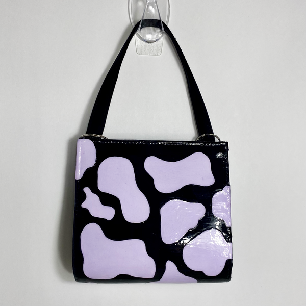 A black purse with pastel purple cow print. The purse is on a white backdrop. 