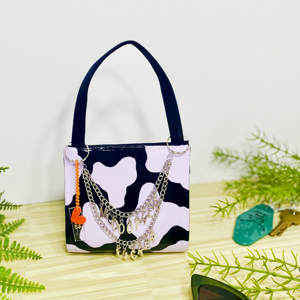 A black purse with pastel purple cow print and draping silver chains with hanging letters that says, "moooother fucker". The purse is on a wood and white backdrop and surrounded by faux plants. 