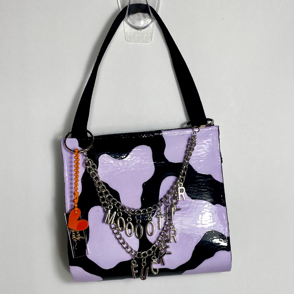 A black purse with pastel purple cow print and draping silver chains with hanging letters that says, "moooother fucker". The purse is on a white backdrop. 