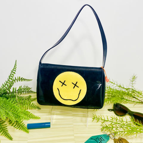 Black handbag with a yellow smiley face in the middle with X's for the eyes and a squiggly smiley face. The purse is surrounded by faux plants on a wooden backdrop.