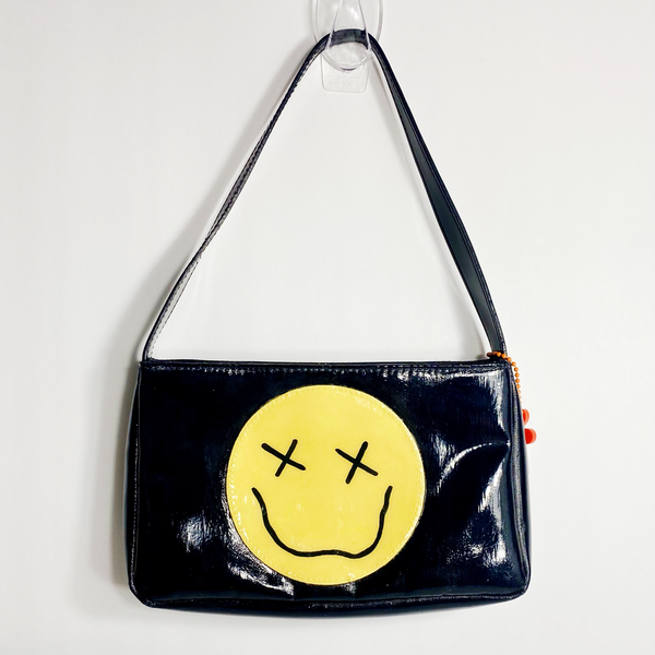 Black handbag with a yellow smiley face in the middle with X's for the eyes and a squiggly smiley face. The purse is on a white backdrop. 