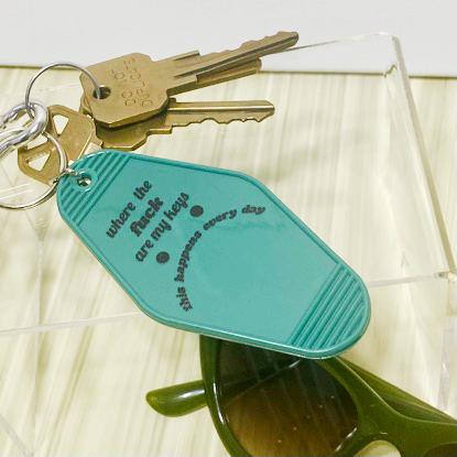 Teal motel style keychain saying "where the fuck are my keys this happens every day," on a wooden and white background. Surround by keys and a pair of green sunglasses.