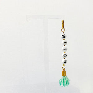 Gold-toned keychain with white acrylic beads with black lettering that spell "witch" with a blue tassel.